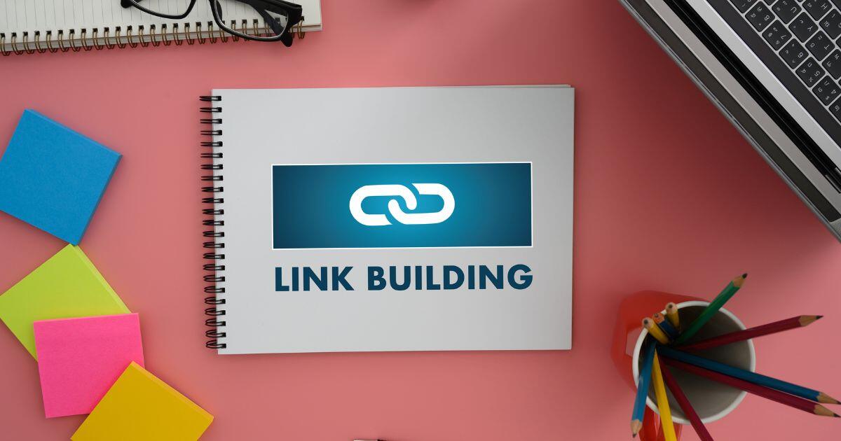 Auf einem rosafarbenen Tisch liegen verschiedene Materialien des Büros. Auf einem Ringblock steht Link Building geschrieben.