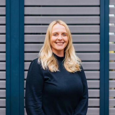 Eine Frau steht vor einer grau gestreiften Wand mit lächelndem Blick in die Kamera. Sie trägt ein schwarzes Kleid und hat blonde, schulterlange Haare.