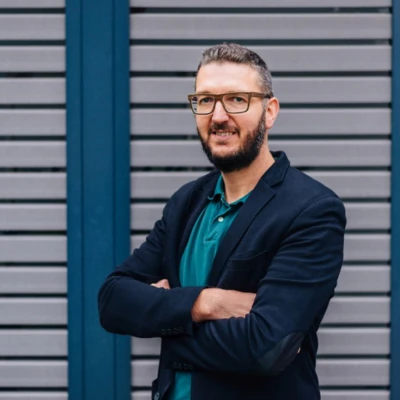 Ein Mann mit dunklen kurzen haaren und Bart steht vor einer grau gestreiften Wand. Er trägt eine Brille, ein grünes Polohemd, ein dunkles Sakko.