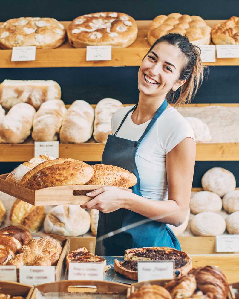 Eine Bäckerin hinter dem Tresen mit weißem T-Shirt und blauer Schürze hält ein Tablet mit frisch gebackenen Broten in den Händen.