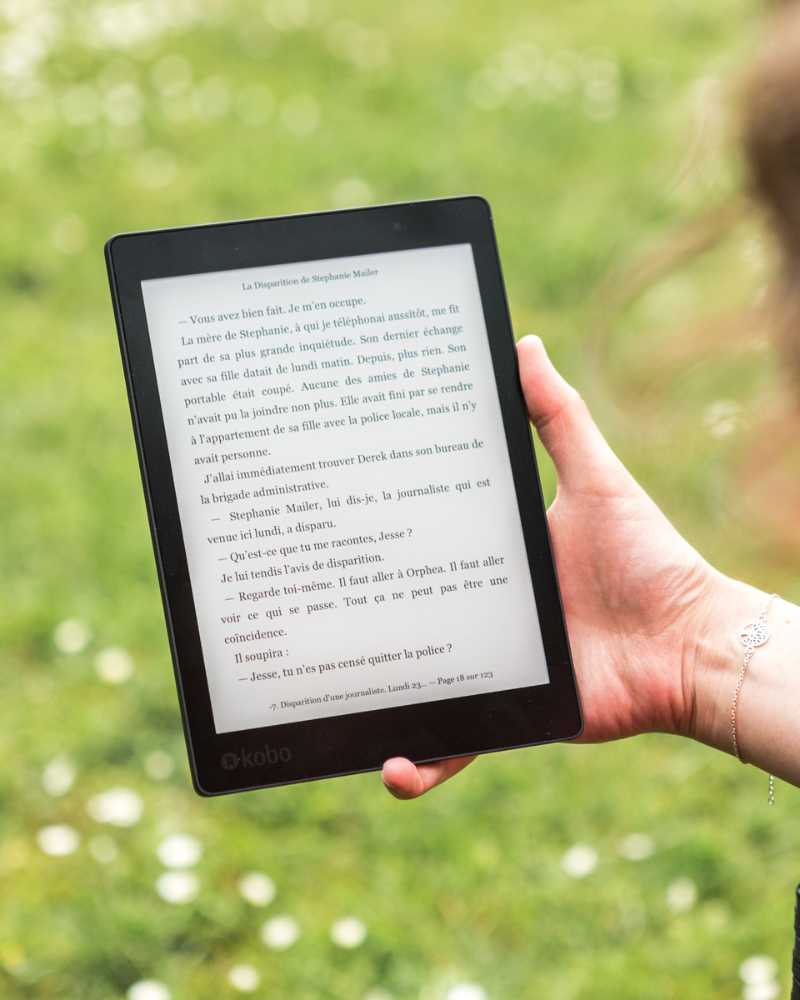 Auf einem E-Book Reader ist französischer Text abgebildet. Eine Person hält das E-Book in der rechten Hand und im Hintergrund ist eine grüne Wiese bei Sonnenschein.