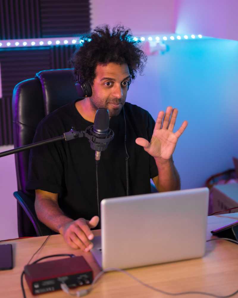 Ein Mann mit schwarzen, lockigen Haaren sitzt während einer Video-Aufzeichnung am Schreibtisch und spricht in ein Podcast-Mikrofon.