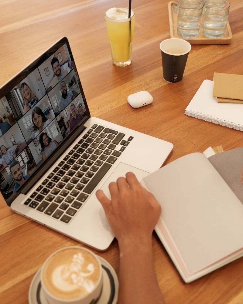 An einem Macbook Bildschirm sind 9 Personen als Teilnehmende eines Webinars dargestellt. Die referierende Person hat einen Milchkaffee auf dem Tisch stehen und Notizbücher daneben liegen.